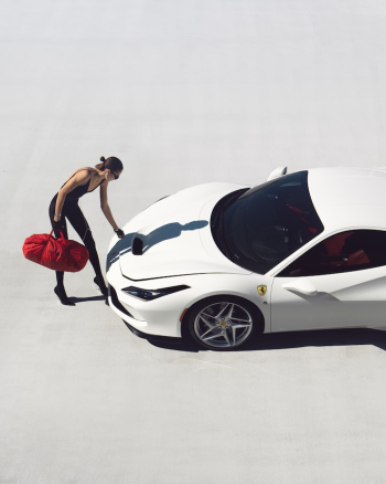 a woman standing next to a white sports car with a red bag on it's hood and her hand on the hood of the car