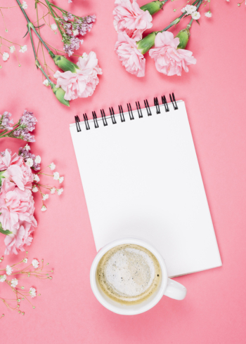 An overhead view of coffee cup on blank notepad with carnations; gypsophila; limonium flowers on pink background