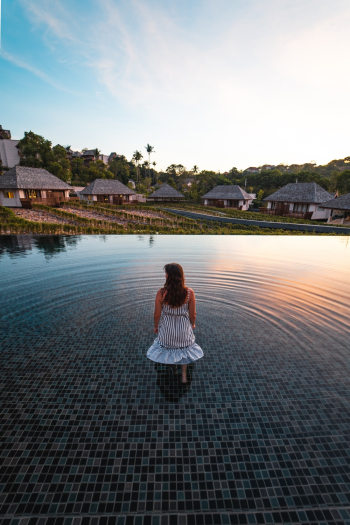 Back View Photo of Woman in White Dress Standing in the Middle of an Infinity Pool Looking at Resort Cottages