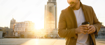 Cropped view of happy man in coat holding coffee to go against sunset on urban street, panorama