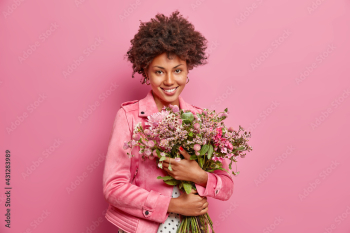 Cute curly haired African American woman embraces big bouquet of flowers recieved from boyfriend has festive mood wears jacket isolated over pink background. Holiday event. Mothers Day concept