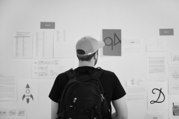 Grayscale Photography of Standing Man Looking on Wall With Pinned Papers
