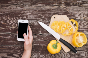 Human hand holding cellphone with slices of yellow bell pepper on chopping board with knife over the wooden desk