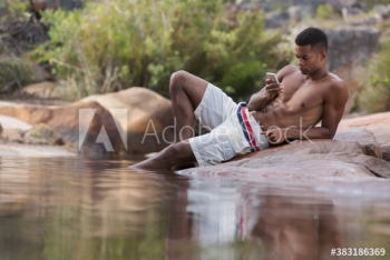 Man in swim trunks using smart phone on rocks at lakeside Man in swim trunks using smart phone on rocks at lakeside
