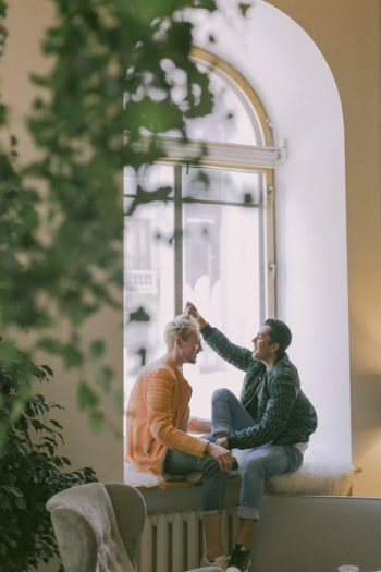 Man Wearing Green Jacket in Front of Man Wearing Orange Zip-up Jacket Sitting on Near the Window Photo