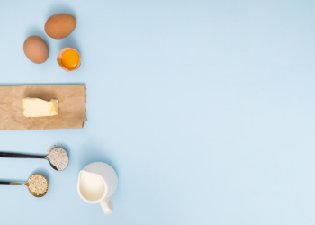 Measuring spoon with oat barn; flour; eggs; butter and milk on blue backdrop