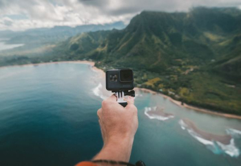 person holding black action camera fronting green mountains person holding black action camera fronting green mountains