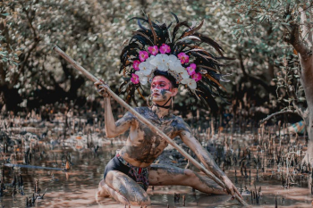 Person Wearing Feather Headdress Holding Wooden Stick While Kneeling on Mud