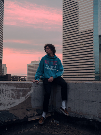 Photo of Man in Hoodie Sitting on Concrete Slab in View of  High-rise Building Under Sky