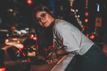 Photo of Smiling Woman In Eyeglasses Leaning on Hand Rail by Sidewalk During Night Time
