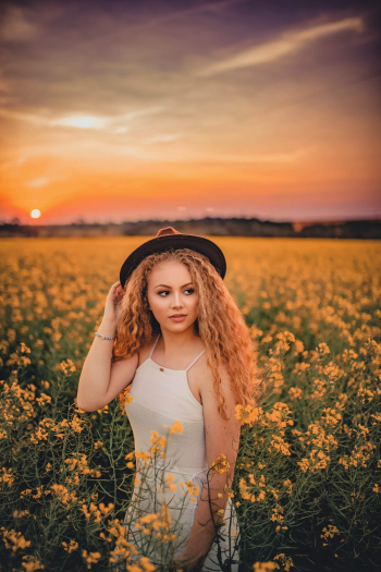 Photo of Woman in White Sleeveless Dress and Brown Hat Standing in Yellow Petaled Flower Field During Golden Hour