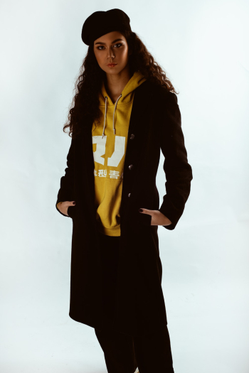 Photo of Woman in Yellow Hoodie, Black Beret Hat, and Black Coat Posing In Front of White Background
