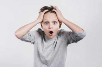 Portrait of a shocked boy with his hands on head looking to camera against grey background