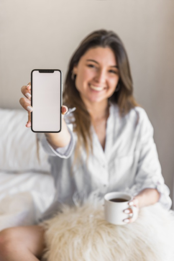 Portrait of a smiling young woman holding coffee cup showing smart phone screen Free Photo