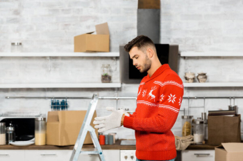Side view of handsome young man putting on working gloves in kitchen with ladder and cardboard boxes during relocation in new home