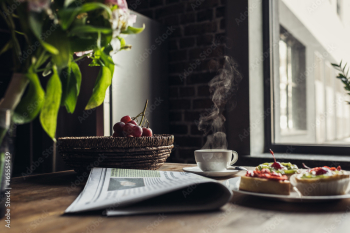 Still life of newspaper, breakfast with cakes and hot coffee on kitchen table in front of window