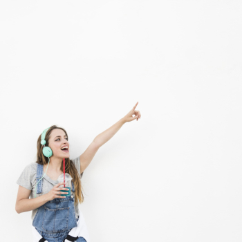 Woman wearing headphone holding glass of juice pointing finger against white backdrop