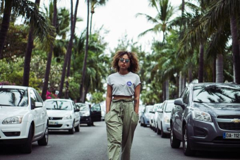 woman with hands on pocket walking between vehicles on road during daytime