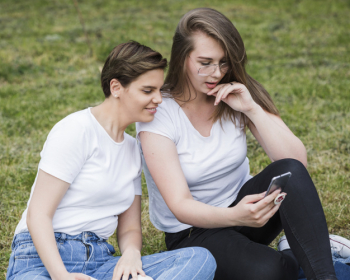 Women friends using smartphone sitting on grass Women friends using smartphone sitting on grass
