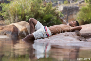 Young man in swim trunks relaxing on rocks at lakeside Young man in swim trunks relaxing on rocks at lakeside