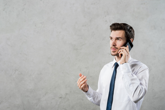 Close-up of a young businessman talking on smartphone looking away against grey concrete wall