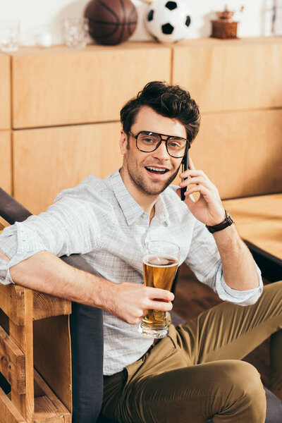 Portrait of cheerful man with glass of beer in hand talking on smartphone in cafe