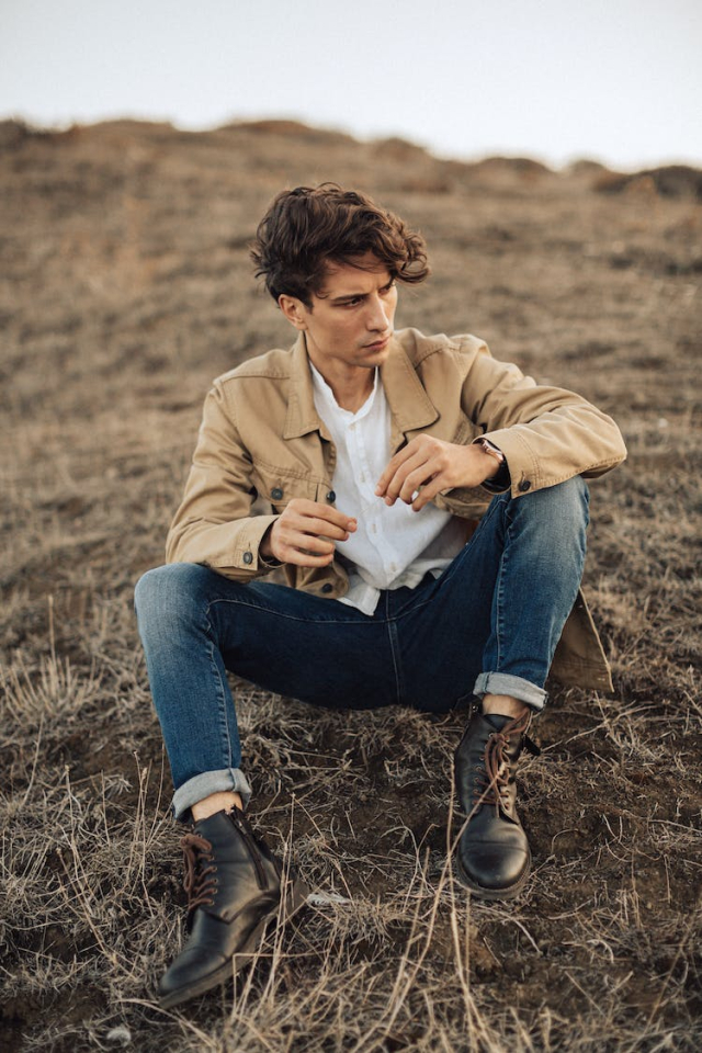 Young Man in Blue Jeans and Beige Jacket Sitting on Ground in Empty Fields